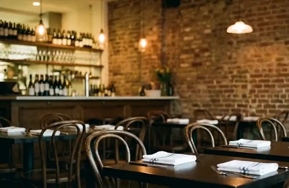 Interior de una trattoria italiana de barrio al atardecer, mesas de madera oscura, ladrillo a la vista y lamparas colgantes encendidas